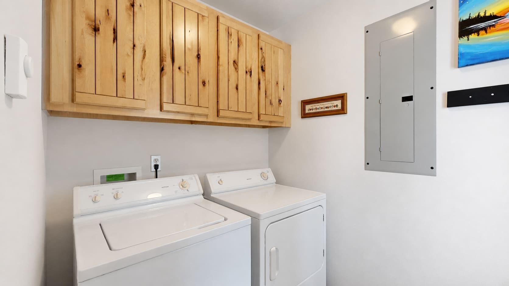 Un cuarto de lavado con dos lavadoras blancas, armarios de madera arriba, un cuadro en la pared y una puerta con pomo.