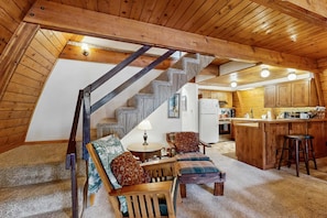 A cozy loft-style room with a wooden staircase, a dining area with a table and chairs, and a kitchen area with wooden cabinets and a bar stool. - Downtown McCall A-Frame Cabin | Walk to Lake (McCall)