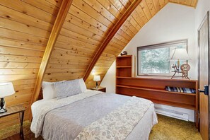 A bedroom with a sloped ceiling, a bed with patterned bedding, a nightstand with a lamp, a window with a view, and built-in shelves. - Downtown McCall A-Frame Cabin | Walk to Lake (McCall)