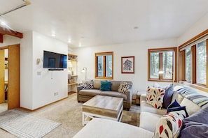 A spacious living room with a flat-screen TV, a sofa set, a coffee table, and large windows offering a view of trees. - Downtown McCall A-Frame Cabin | Walk to Lake (McCall)