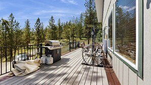 Une terrasse en bois équipée d’un barbecue, de mobilier d’extérieur et d’une grande fenêtre donnant sur une forêt.