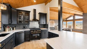 A modern kitchen with dark cabinetry, a central stove, and large windows offering a view of the outdoors. - Luxury Mountain View Retreat | Hot Tub & Game Room (McCall)