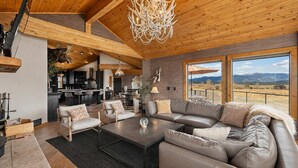 A spacious living area with a large sofa, armchairs, and a coffee table. There is a kitchen in the background, a chandelier, and a view of mountains through large windows. - Luxury Mountain View Retreat | Hot Tub & Game Room (McCall)