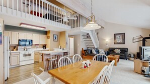 Ampla sala de estar com mesa de jantar de madeira, cadeiras brancas, sofá e televisão. É visível uma cozinha com armários brancos e micro-ondas ao fundo.