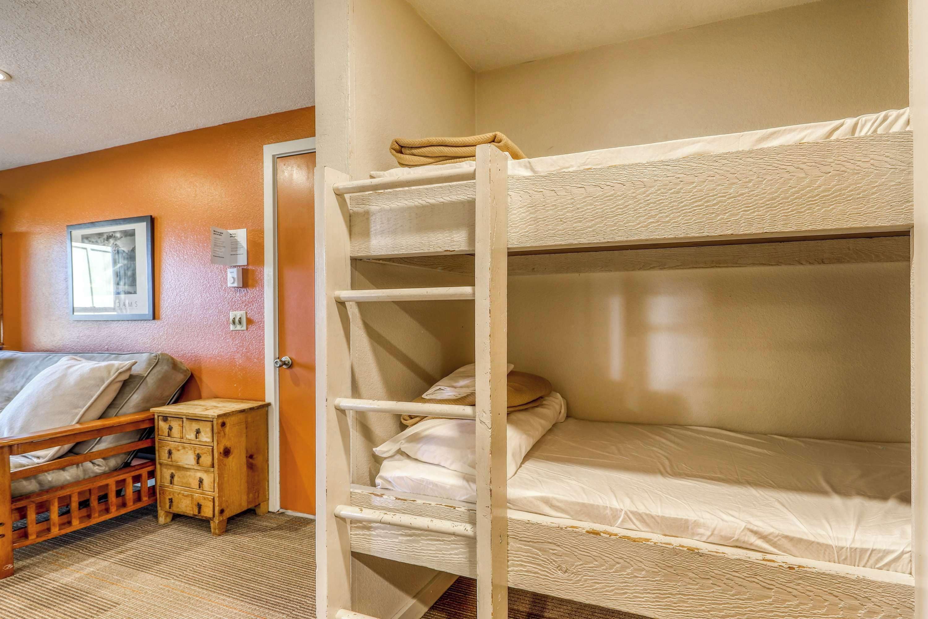 Una cama individual superpuesta con una escalera de madera, una mesita de noche de madera con cajones, y un cuadro enmarcado en la pared.