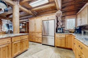 A wooden cabin interior with a stone floor, wooden cabinets, a stainless steel refrigerator, and a dining area with a table and chairs. - Luxury McCall Log Cabin Hot Tub & Gourmet Kitchen (McCall)
