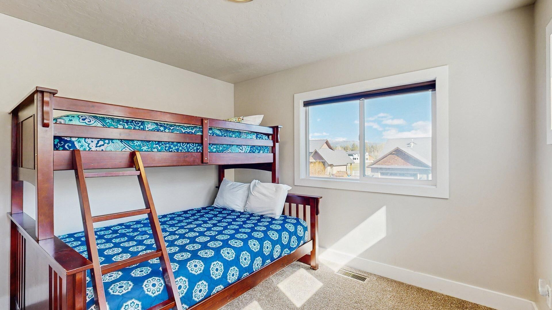A bunk bed with a ladder, a window with a view of houses, and a carpeted floor.