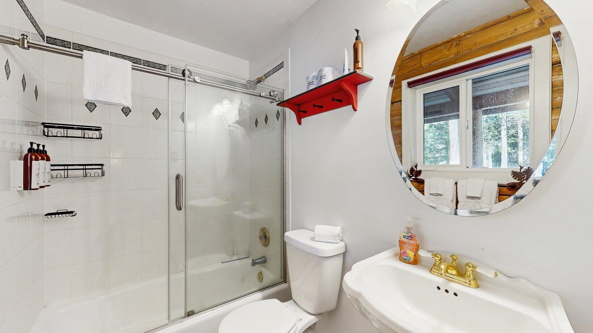 A bathroom with a glass shower enclosure, a toilet, a sink with a gold faucet, a mirror with a wooden frame, and a window with a view of trees.