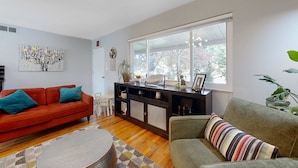 Un salon avec un canapé rouge, un fauteuil vert, un plancher en bois, une grande fenêtre et un meuble de rangement noir pour appareils électroniques.