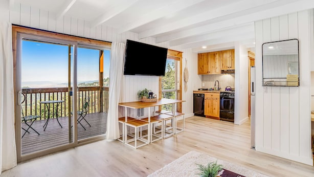 A spacious living area with a wooden table and chairs, a flat-screen TV, and a sliding glass door leading to a balcony with a view of the outdoors.