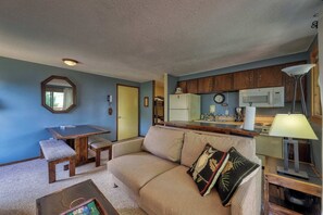 A living room with a sofa, a wooden table with a bench, a kitchen area with wooden cabinets and a microwave, and a lamp on a side table. - Slope-Side Getaway Deck Pool Table Views (Boise)