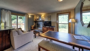 A cozy living area with a sofa, a wooden table, and a TV. There is a fireplace and a window with a view of greenery.