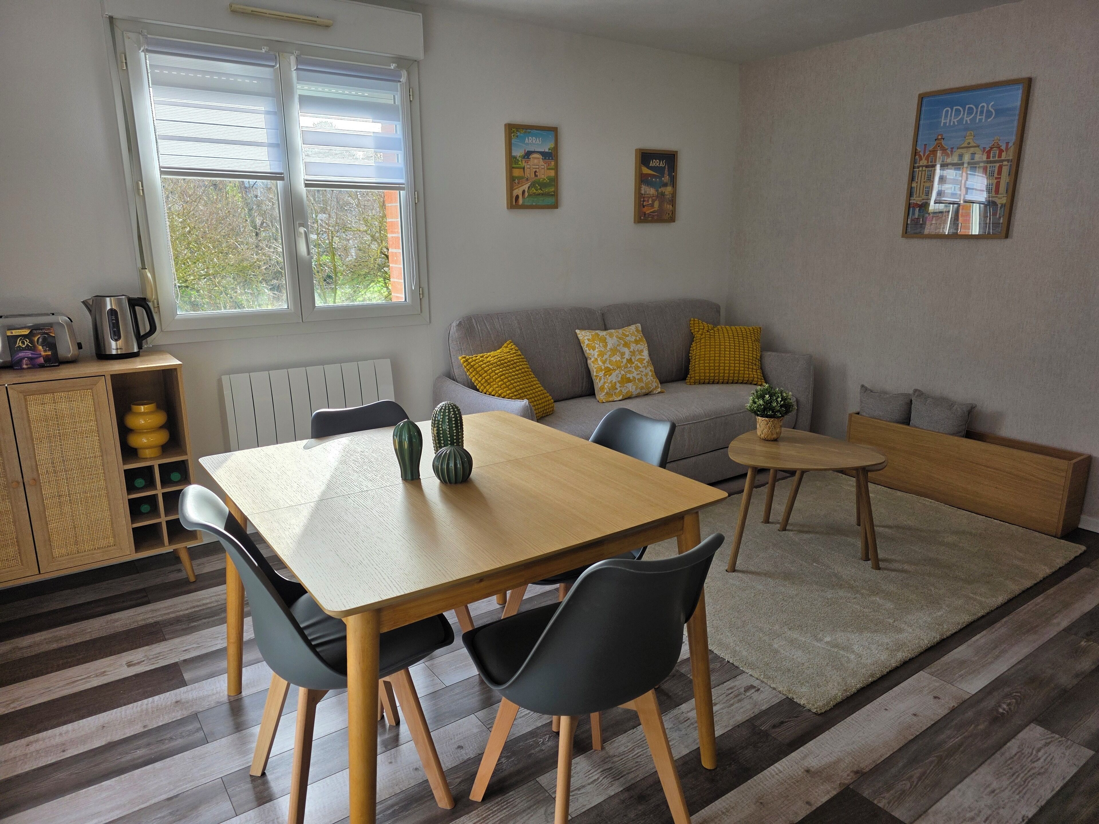 A modern living room with a wooden dining table, two black chairs, a grey sofa with yellow cushions, and a small round table with a plant.