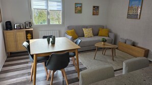 Ein gemütliches Wohnzimmer mit einer Couch, einem Esstisch und einem Couchtisch. An den Wänden hängen zwei Bilder, ein Heizkörper und ein Fenster mit Jalousien.