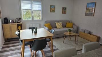 Een gezellige woonkamer met een bank, een eettafel en een salontafel. Twee schilderijen hangen aan de muur, er is een radiator en een raam met jaloezieën.