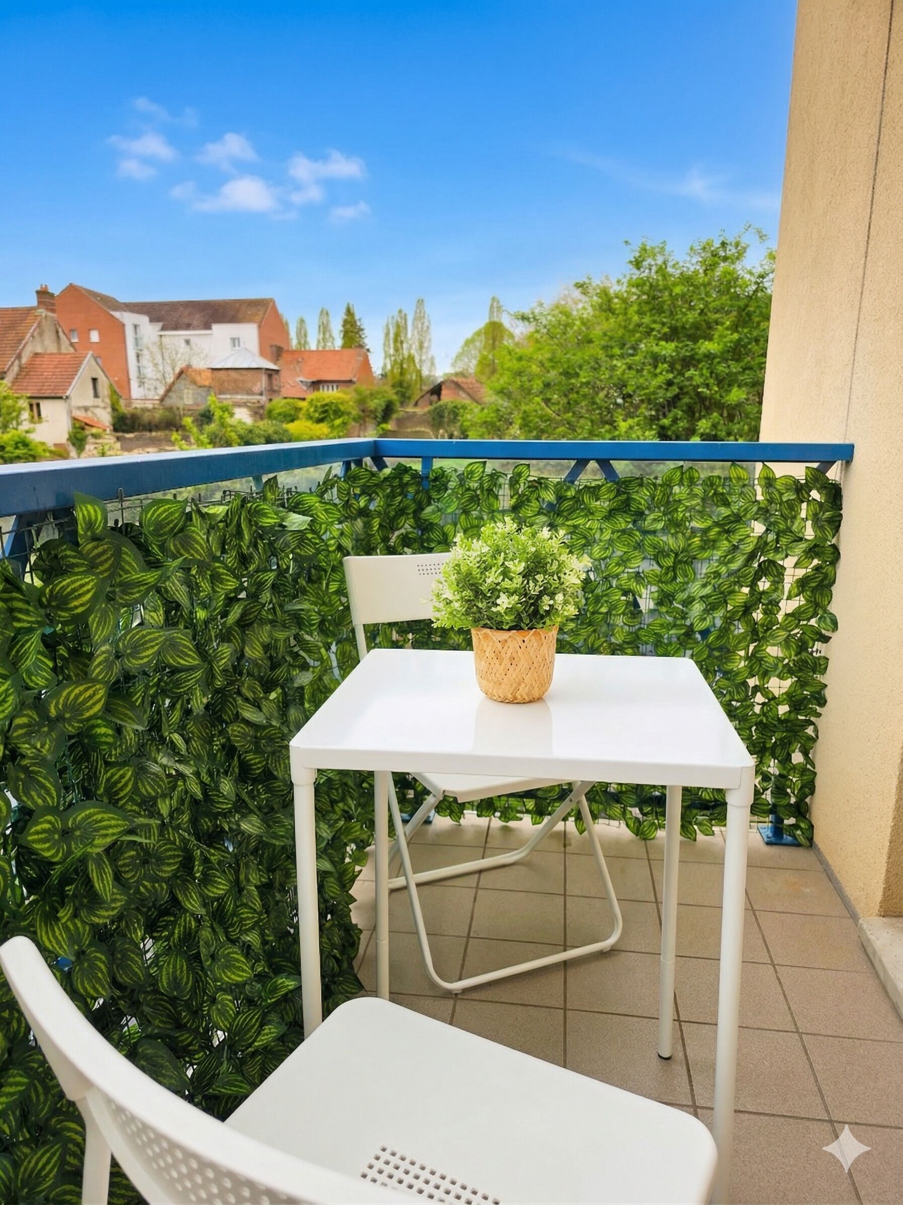 A balcony with a white table and chair, a potted plant, and a view of houses and trees.
