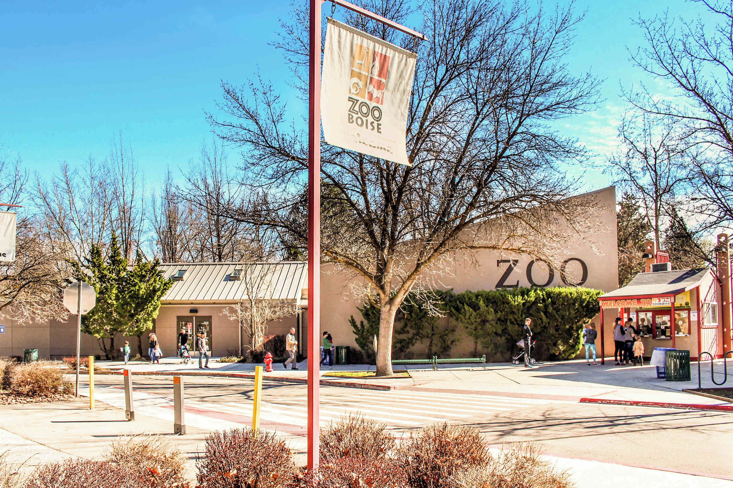 A zoo entrance with a large sign, a building with a 'ZOO' sign, and people walking around.