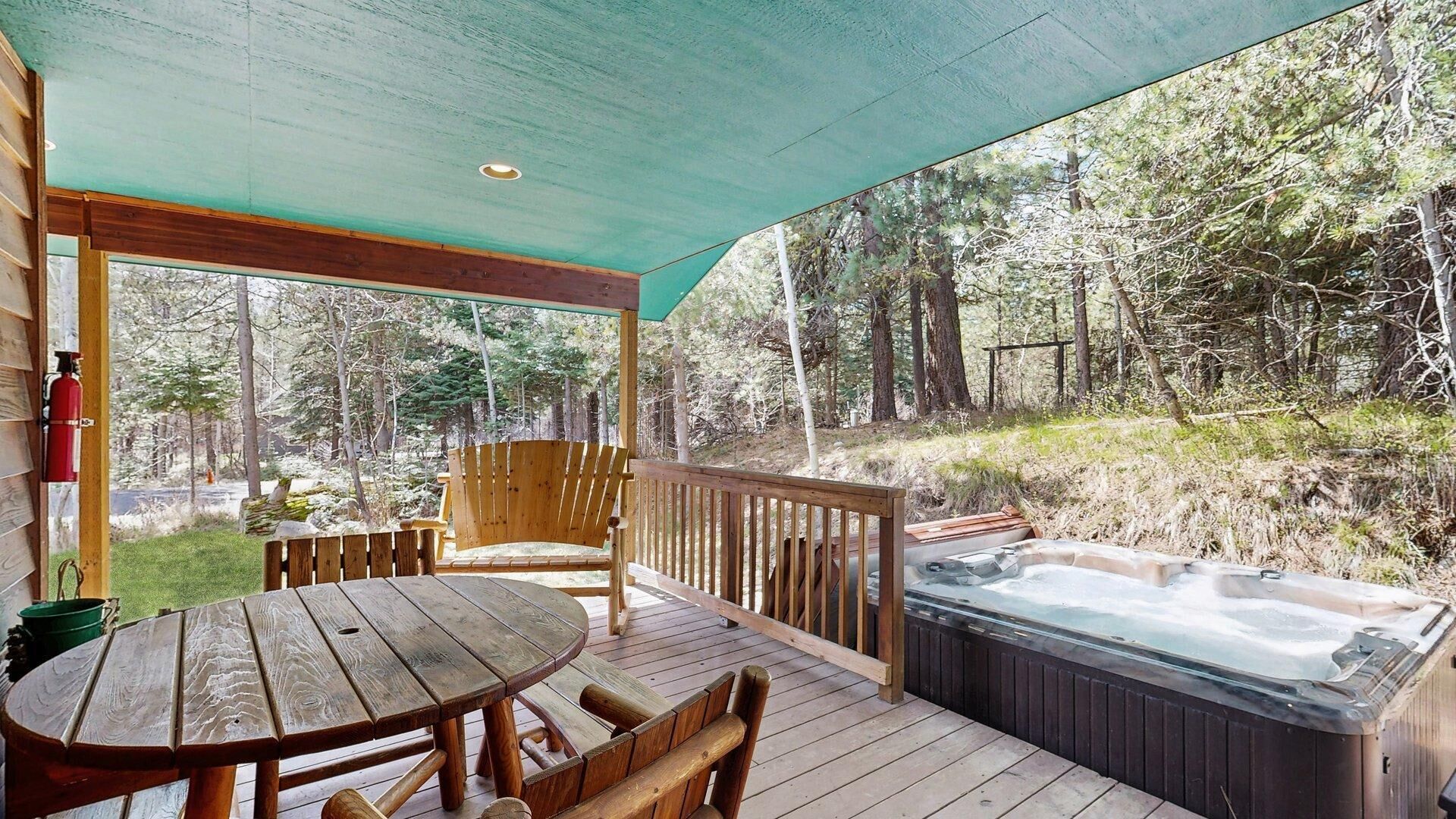 Una terraza de madera con un jacuzzi, una mesa y sillas, rodeada de bosque.