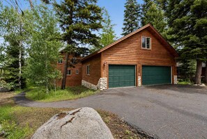 A wooden cabin with two green garage doors, surrounded by trees. - Luxury McCall Home | Views, Game Room, Deck, Pool (McCall)
