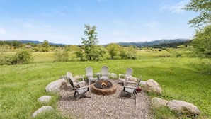 A fire pit with four Adirondack chairs arranged around it in a grassy field with mountains in the background.