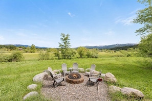 A fire pit with four Adirondack chairs arranged around it in a grassy field with mountains in the background. - Luxury McCall Home | Views, Game Room, Deck, Pool (McCall)