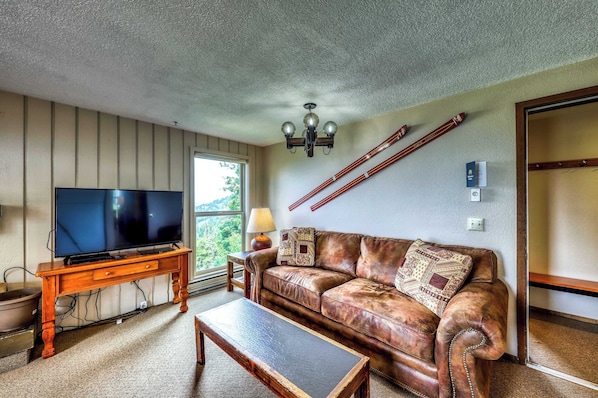 Ein Wohnzimmer mit Fernseher, Couchtisch aus Holz, Ledersofa und einem großen Fenster.