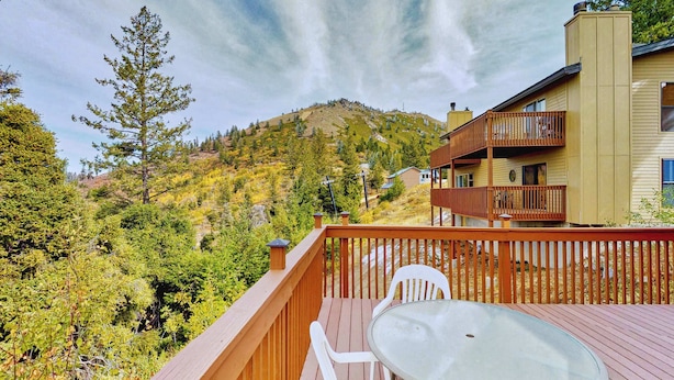 A deck with a glass table and chairs, overlooking a forested hillside and a building with a balcony. - Ski-In Ski-Out Bogus Basin Condo with 2 Decks (Boise)