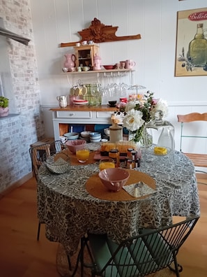 Un coin repas avec une table ronde recouverte d’une nappe à motifs, entourée de chaises. Une étagère en bois, située au-dessus, contient divers ustensiles de cuisine et de la verrerie.