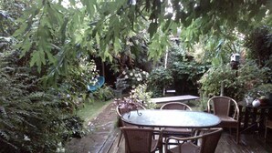 Une terrasse extérieure avec une table ronde et des chaises en osier, entourée d’une végétation luxuriante et bordée d’un chemin en pierre.