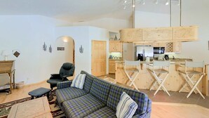 A living room with a sofa, a chair, and a small table. There is a kitchen area with wooden cabinets and a bar counter. - McCall Golf Home | Patio, Fenced Yard & Views (McCall)