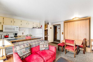 A compact living space with a kitchenette, a red sofa with patterned cushions, a wooden dining table, and a red chair.