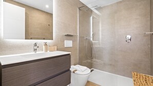 Et moderne badeværelse med et stort spejl, en hvid vask, et mørkt trækommode og en brusekabine i glas samt et hvidt toilet.