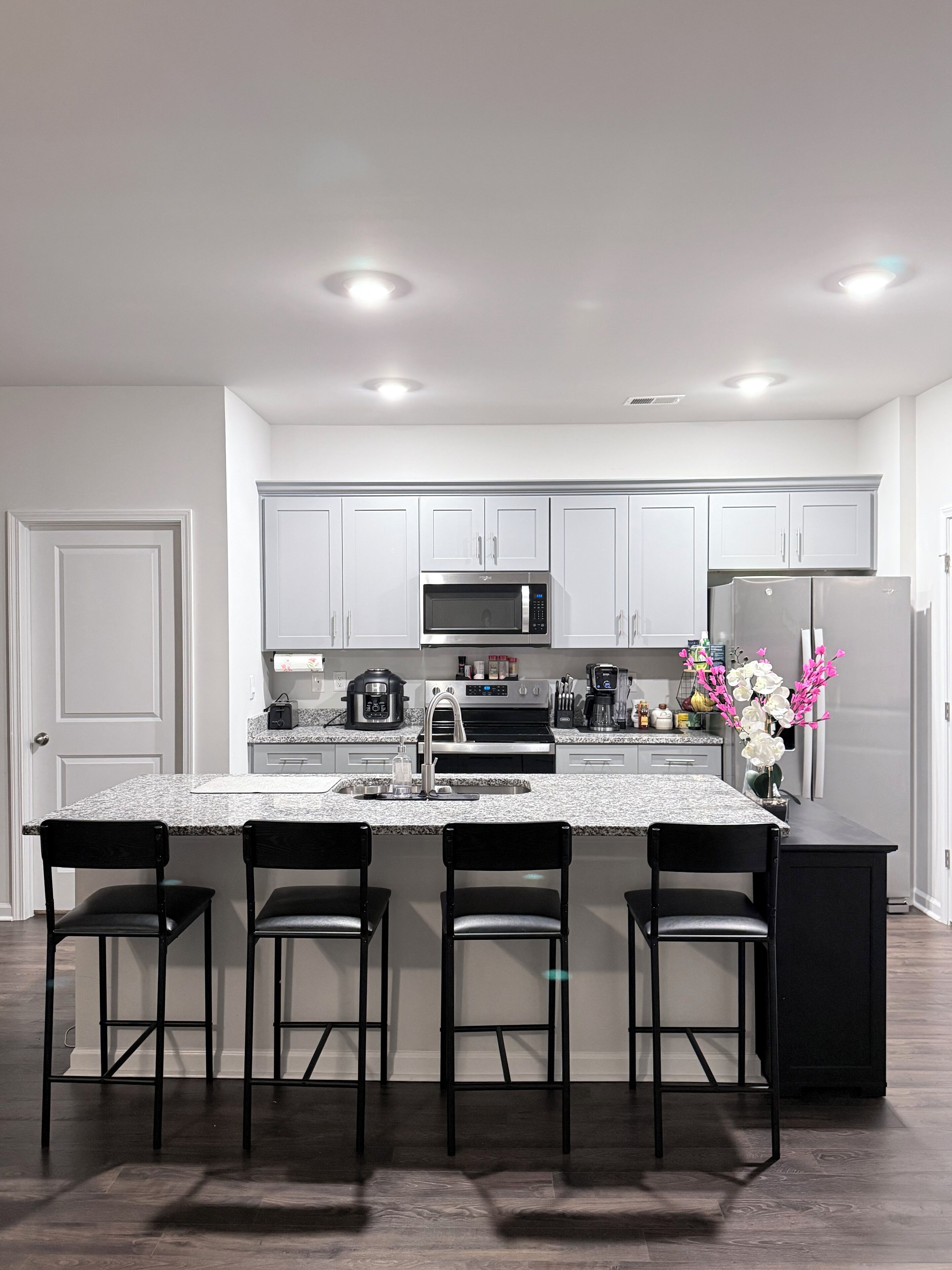 A modern kitchen with white cabinets, a granite countertop, a stainless steel refrigerator, a microwave, and a coffee maker. There are four black bar stools at the island.