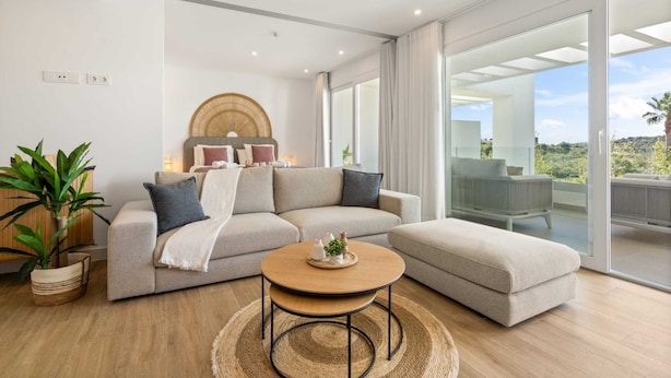 Ein modernes Wohnzimmer mit Sofa, Hocker und Couchtisch, einem großen Fenster mit Aussicht und einem Topfpflanzen.