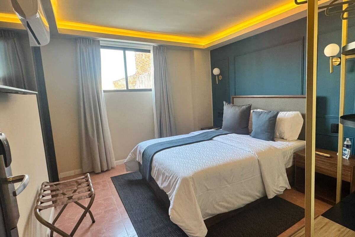 A hotel room with a bed, a small table, a chair, a window with curtains, and a wall-mounted lamp.