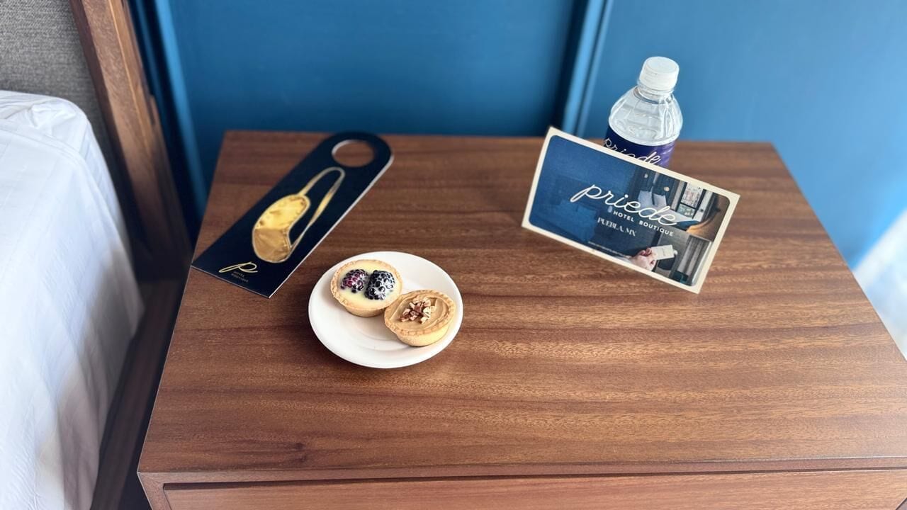 A wooden nightstand with a box of pastries, a plate with two pastries, a bottle of water, and a brochure.