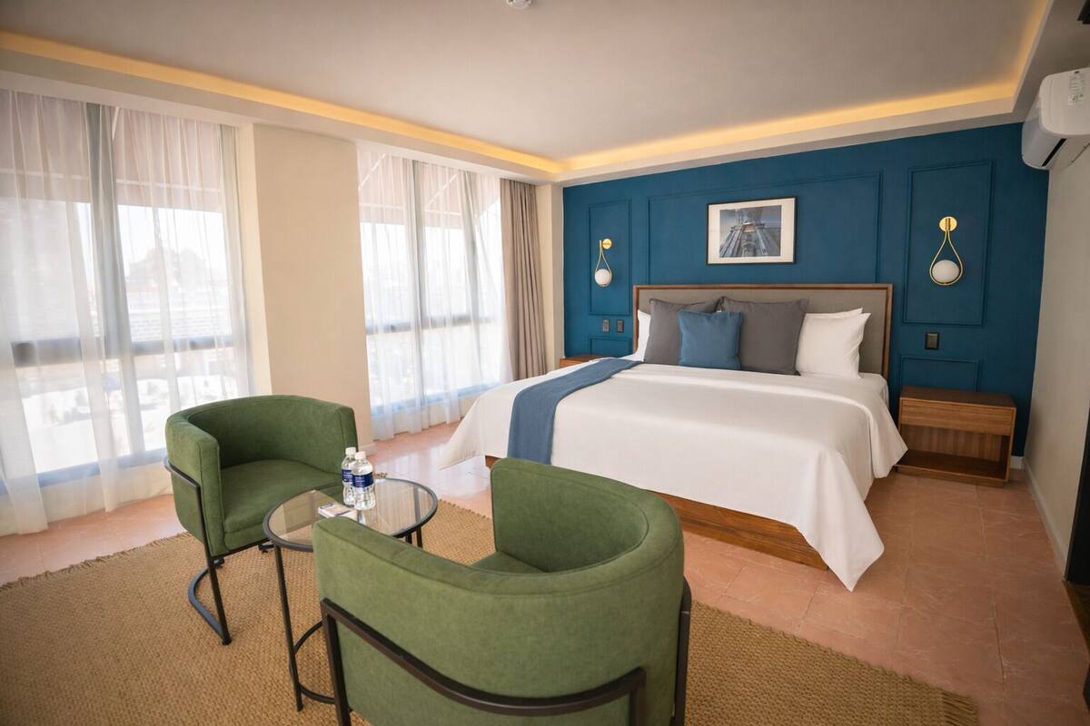 A hotel room with a bed, two green armchairs, a small table with water bottles, and a large window with sheer curtains.