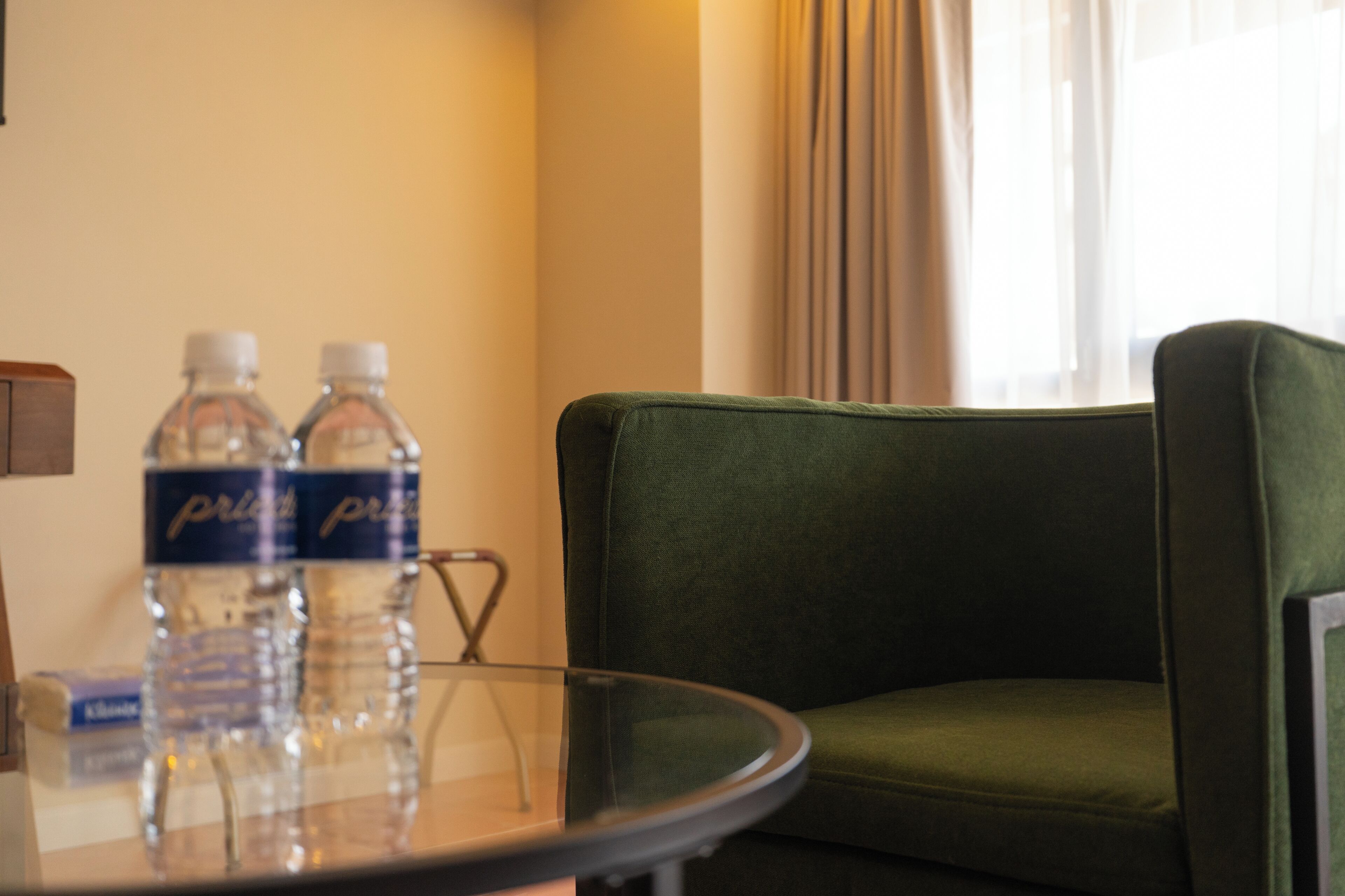 A hotel room with a green sofa, a glass table with water bottles, and a window with curtains.