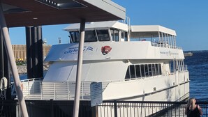 A ferry with a white exterior and a logo on the side, docked at a pier with a metal railing and a sign indicating no skateboarding. - Sand Dollar 6mi-Pensacola Bch (Gulf Breeze)