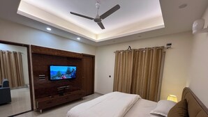 A modern hotel room with a large bed, a flat-screen TV mounted on a wooden cabinet, a ceiling fan, and a large window with curtains.