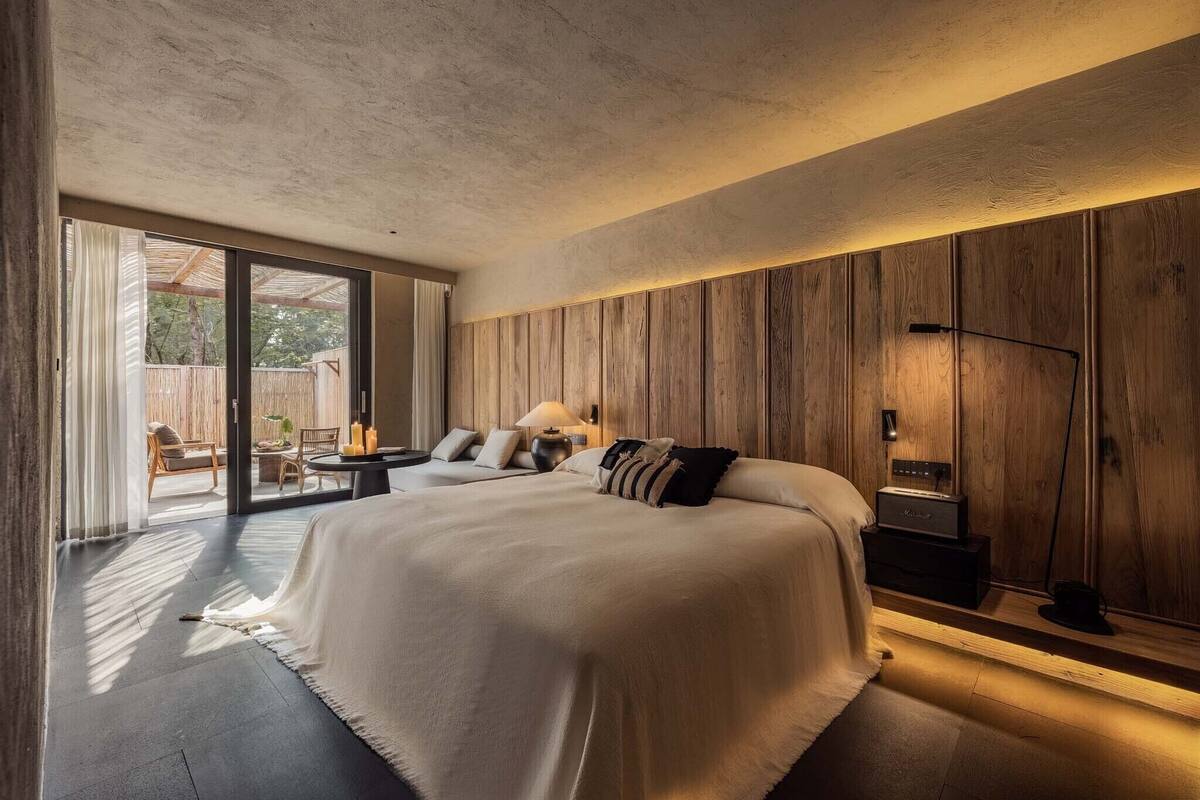 A modern bedroom with a large bed, wooden panel walls, a small seating area, and a view through a sliding door.