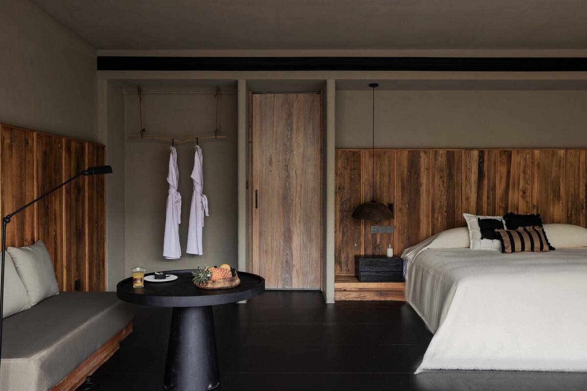A modern hotel room with a large bed, a sofa, a round table with a drink and fruit, and wooden paneling on the walls.