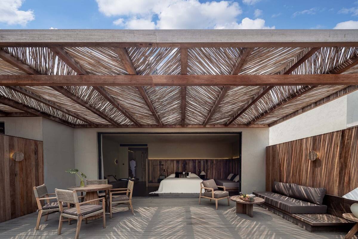 A covered outdoor area with a wooden deck, seating arrangements, and a view into a room with a bed and a bathroom.
