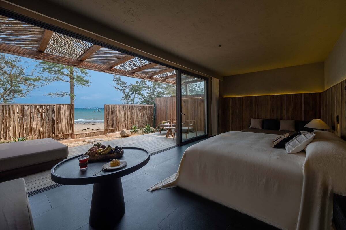 A bedroom with a large bed, a small table with fruit, and a view of the beach.