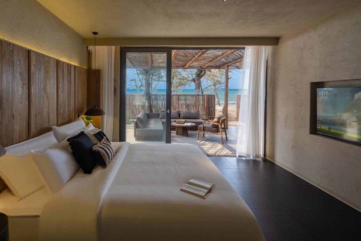 A modern hotel room with a large bed, a view of the beach, and a dining area with outdoor furniture.