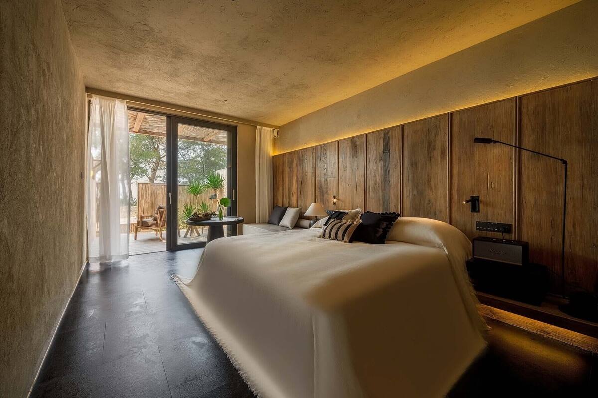 A modern bedroom with a large bed, wooden paneling on the walls, a desk with a lamp, and a view through a glass door to an outdoor area.