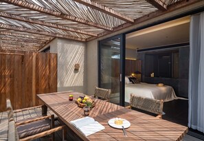 A modern outdoor dining area with a wooden table, wicker chairs, and a glass sliding door leading to a bedroom with a bed and a nightstand. - Weizhou Island Coral Reef Hotel (Beihai)