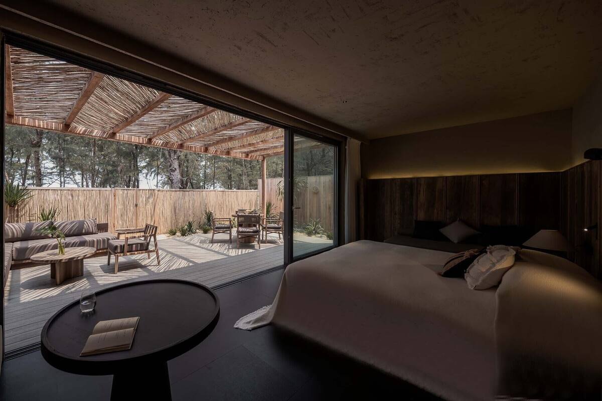 A modern bedroom with a large bed, a small table, and a view of an outdoor patio with seating and a wooden fence.
