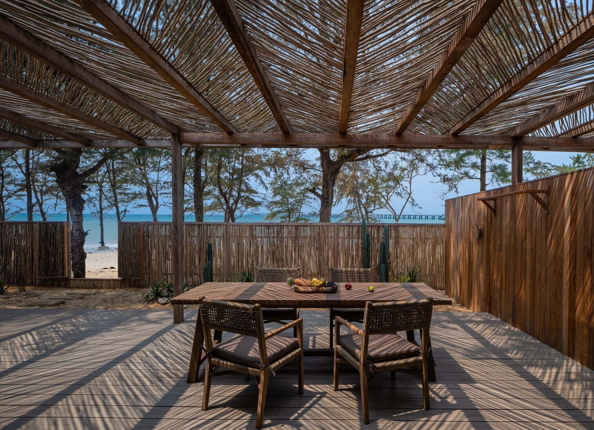 Een terras buiten met een houten tafel en stoelen, een geweven plafond en uitzicht op het strand en de oceaan.