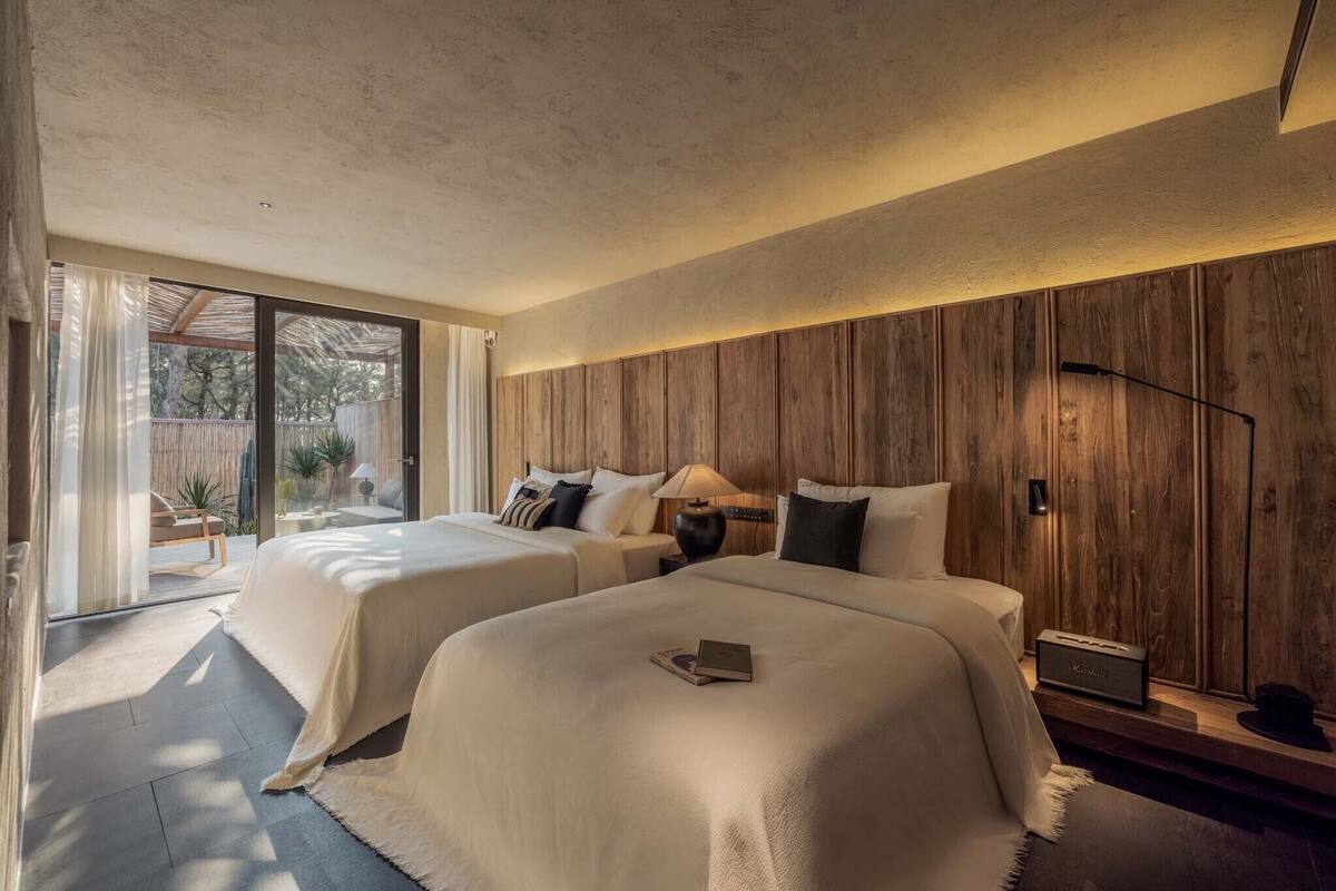 A modern bedroom with two beds, a wooden wall panel, a nightstand, and a lamp.